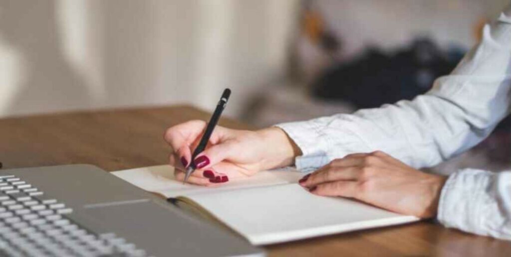 Person writing film pre-production checklist and workflow notes beside laptop