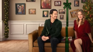Merry Little Ex-Mas Alicia Silverstone and Pierson Fodé standing in a kitchen behind a table covered with gingerbread house supplies; Silverstone looks concerned while Fodé leans on the counter wearing a festive Christmas sweater.