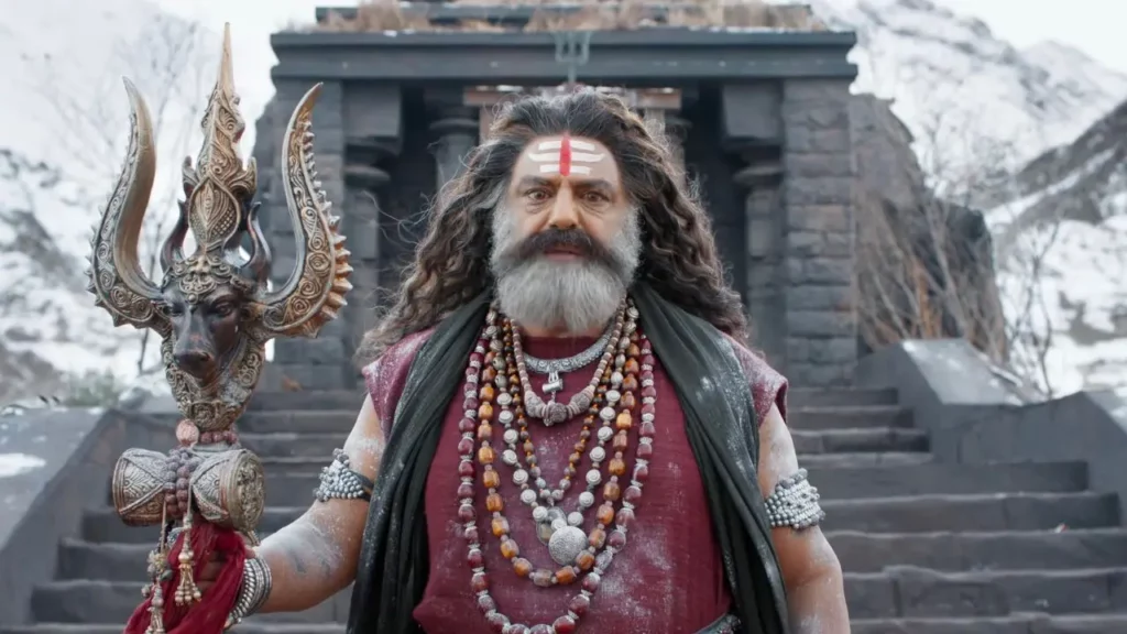 Balakrishna in a traditional Aghora look wearing Rudraksha beads and holding a Trishul in front of a snowy Himalayan temple in Akhanda 2.