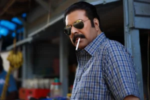 A close-up shot of actor Mammootty in character for the movie 'Kalamkaval', wearing sunglasses and a checkered shirt, with a cigarette in his mouth, looking intensely towards the side.
