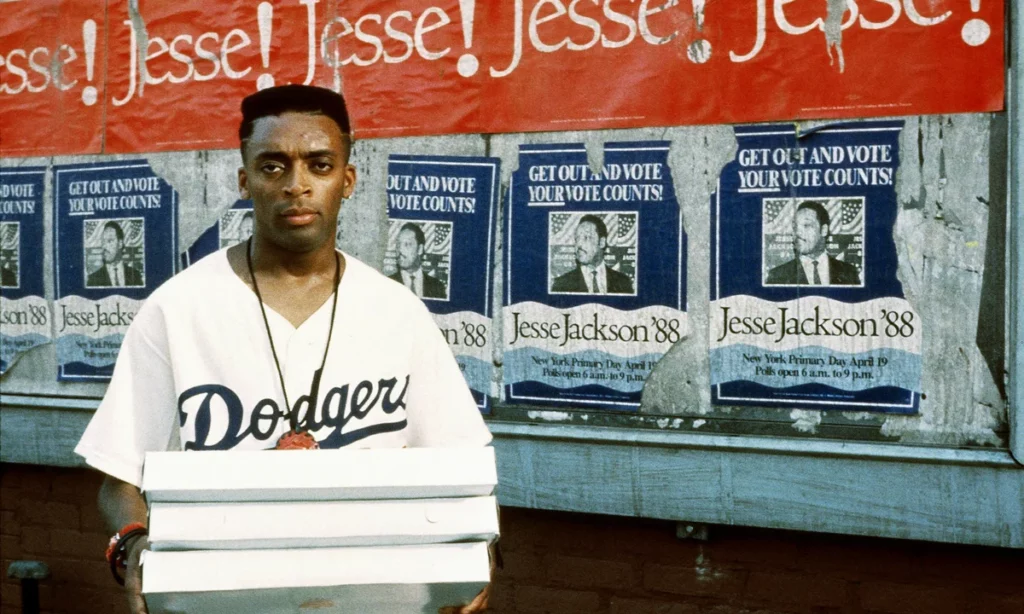 Spike Lee's character Mookie holding pizza boxes in front of a wall covered in Jesse Jackson 1988 campaign posters in the movie Do the Right Thing.