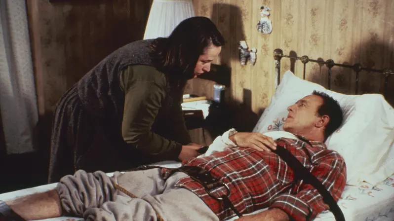 Kathy Bates leaning over a bedridden James Caan, who is strapped to the bed, in a tense scene from the thriller Misery.