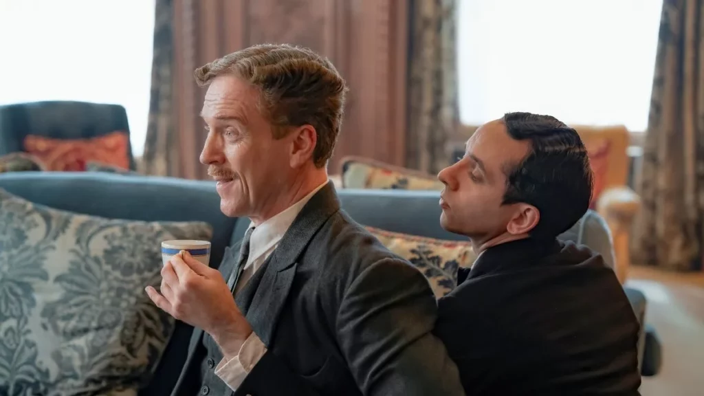Damian Lewis holding a teacup while a servant character stands awkwardly close behind him in a humorous interior scene from the spoof comedy Fackham Hall.