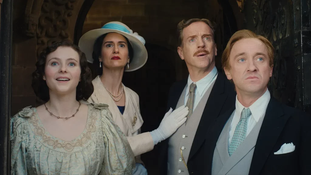 Thomasin McKenzie, Katherine Waterston, Damian Lewis, and Tom Felton standing in a stone archway looking upward with surprised expressions in the movie Fackham Hall.