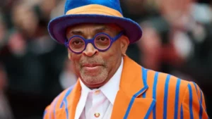 A close-up portrait of director Spike Lee wearing a bright orange and blue striped suit, matching fedora, and colorful glasses on a red carpet.
