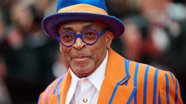 A close-up portrait of director Spike Lee wearing a bright orange and blue striped suit, matching fedora, and colorful glasses on a red carpet.