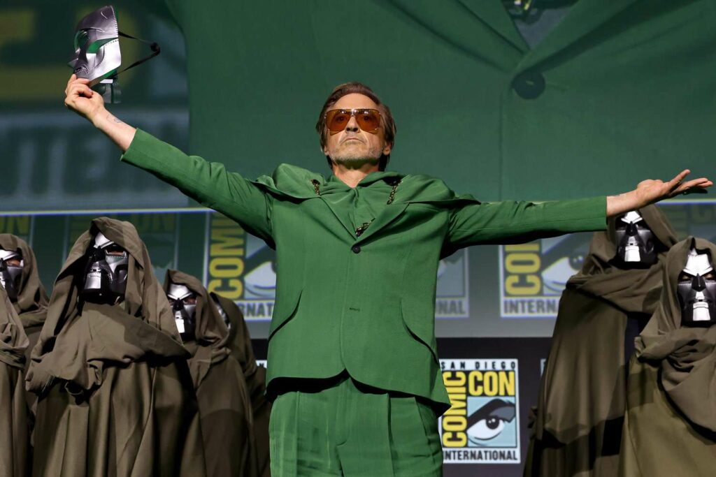 Robert Downey Jr. standing on stage at San Diego Comic-Con wearing a green suit and holding the Doctor Doom mask, announcing his role in the Avengers Doomsday production.