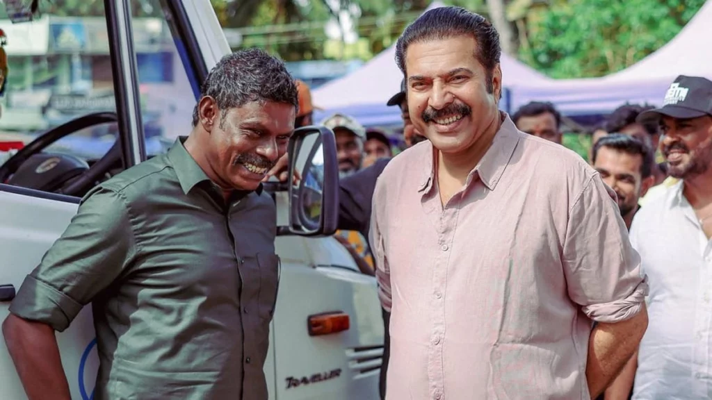 A candid behind-the-scenes photo from the set of 'Kalamkaval' showing actors Mammootty and Vinayakan sharing a laugh near a white van, with crew members visible in the background.