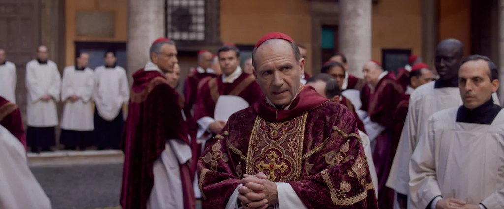 Ralph Fiennes dressed as Cardinal Lawrence standing in a courtyard in the movie Conclave.