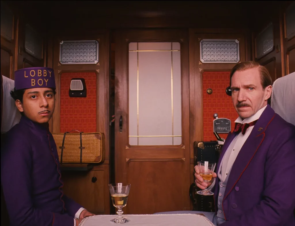 Zero and M. Gustave from The Grand Budapest Hotel wearing signature purple uniforms in a train carriage, showcasing Wes Anderson's pastel color palette.