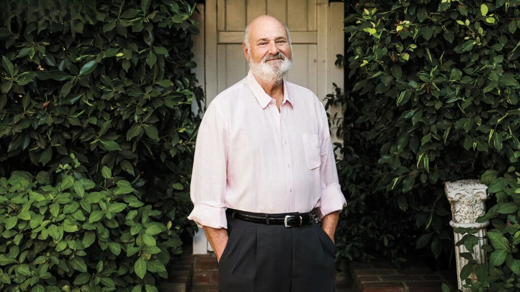 A portrait of film director Rob Reiner standing outdoors in front of lush greenery, wearing a light pink button-down shirt.