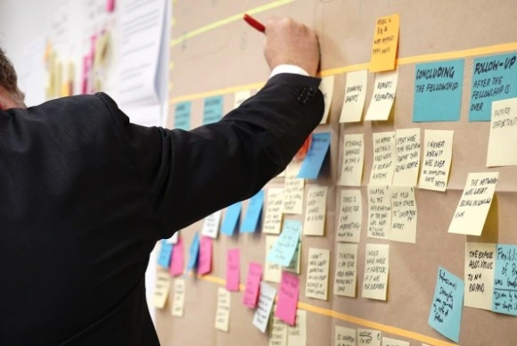 A close-up shot of a filmmaker in a suit pointing to a large brown board covered in dozens of color-coded sticky notes and documents, illustrating a detailed manual shot listing and production planning process.