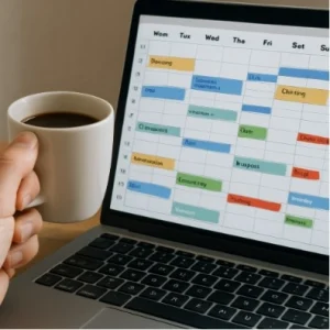 A close-up of a human hand holding a coffee cup next to a laptop screen displaying a complex, organized calendar.