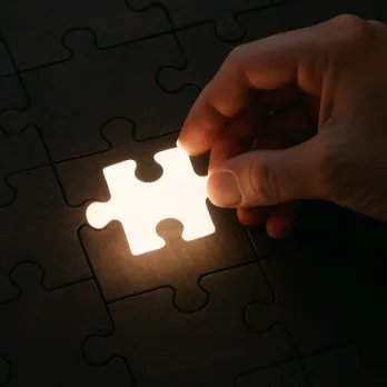 A single hand placing a glowing, white puzzle piece into a dark wooden jigsaw puzzle.