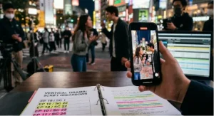 A close-up of a hand holding a smartphone recording a vertical video format scene. In the blurry background, a man and woman act out an intense conversation on a busy city street at night. In the foreground, an open binder titled "Vertical Drama: Script Breakdown" lies on a wooden table alongside a laptop screen displaying production schedules.